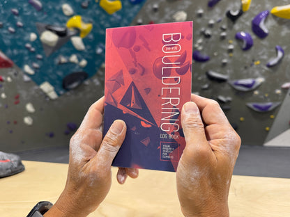 Climber's chalky hands holding Bouldering Log Book" in front of bouldering wall at climbing gym.