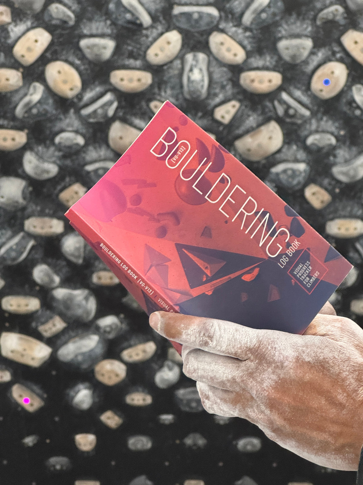 Chalky hands of climber holding "Bouldering Log Book" in front of training board.