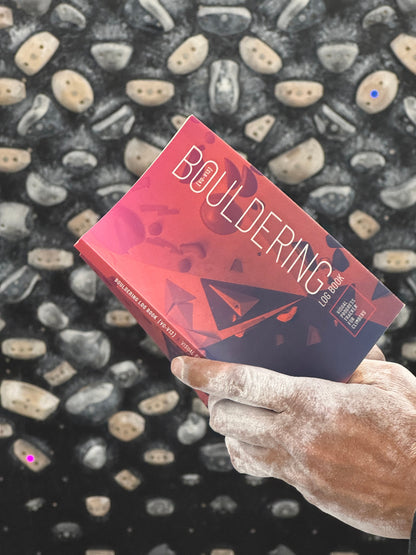Chalky hands of climber holding "Bouldering Log Book" in front of training board.